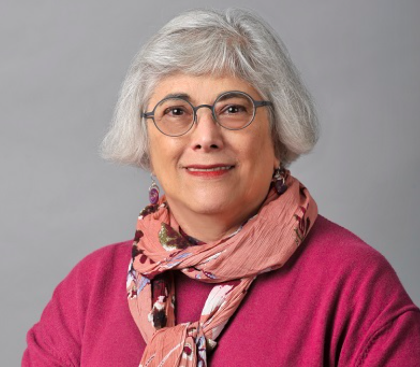 Headshot of a School of Social Work faculty member with short gray hair and glasses, wearing a bright pink top and a beaded necklace, smiling against a neutral background.